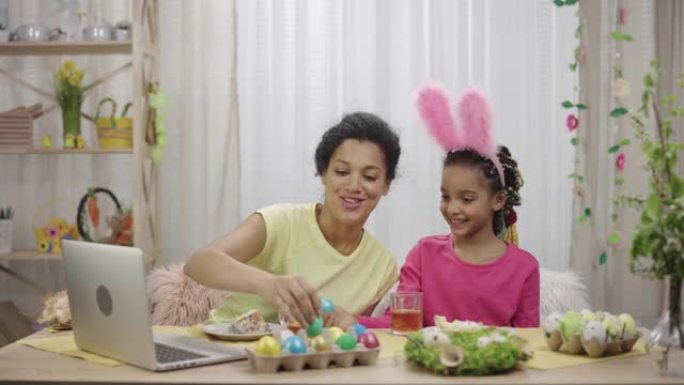 妈妈和女儿通过笔记本电脑进行视频通话，打鸡蛋和复活节蛋糕喝茶。非裔美国妇女和小女孩坐在装饰的房间里。