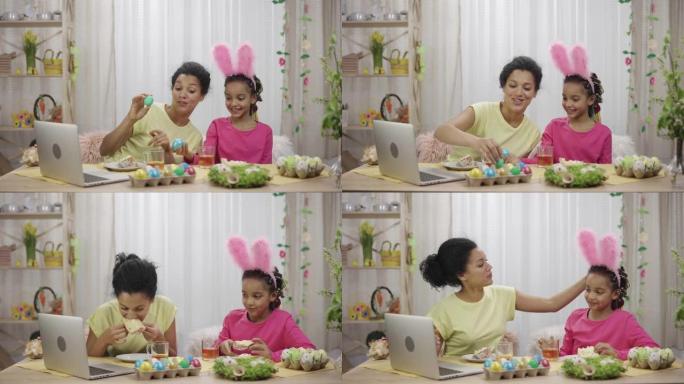 妈妈和女儿通过笔记本电脑进行视频通话，打鸡蛋和复活节蛋糕喝茶。非裔美国妇女和小女孩坐在装饰的房间里。