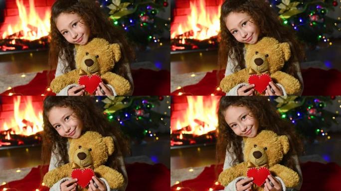 可爱的女孩，卷发微笑着坐在红色格子上，拿着一只泰迪熊和一颗红色的心在壁炉的背景下