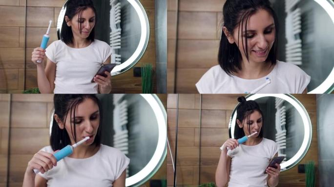 可爱的快乐年轻黑发女人站在镜子旁，用牙刷清洁牙齿，同时在电话上观看有趣的视频