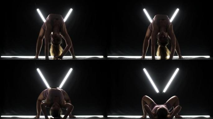 练习瑜伽姿势的健康女性。剪影女孩在黑色背景下的工作室里做运动，带v形白色发光二极管灯管。无压力内部平