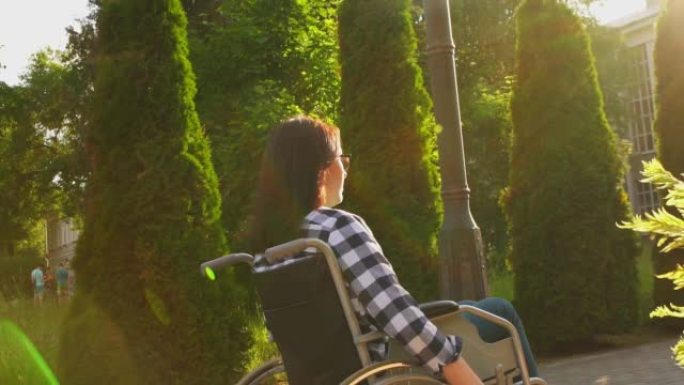 一个残疾女孩穿着一件衬衫，坐在轮椅上，在日落时穿过公园。太阳眩光。缓慢的莫