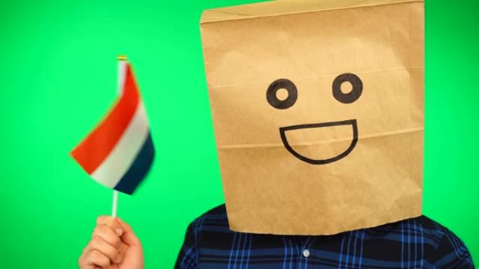头上戴着纸袋的男子的肖像，在绿色的背景下挥舞着荷兰国旗，面带微笑。