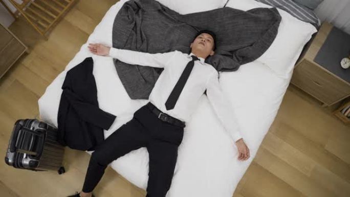 从上面看慢动作不堪重负的亚洲男经理正躺在床上，双臂张开，在家里的卧室里放着一个手提箱。