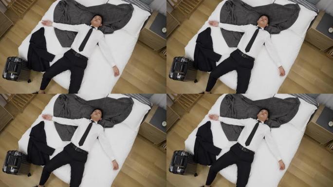 从上面看慢动作不堪重负的亚洲男经理正躺在床上，双臂张开，在家里的卧室里放着一个手提箱。
