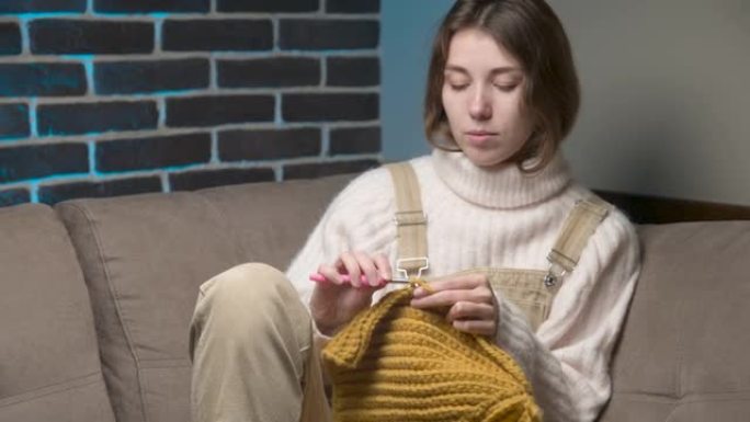 年轻的白人妇女正坐在家里的沙发上编织羊毛制品。国内生产羊毛服装