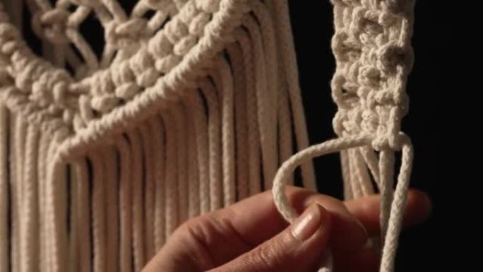 女人用自己的手编织花边线的图案，用于装饰或家居装饰。在黑色背景的创意工作坊中，女人的手在绳子上打结。