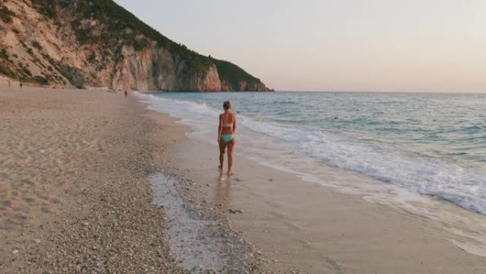 空中低空跟随成年女性欣赏希腊爱奥尼亚岛莱夫卡达美丽的米洛斯海滩的景色。日落金色的阳光和原始的海浪打破