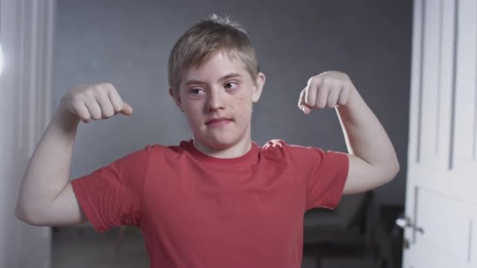 唐氏综合症男子的肖像。这孩子在家展示肌肉和二头肌。家庭锻炼和运动。有特殊需要的人