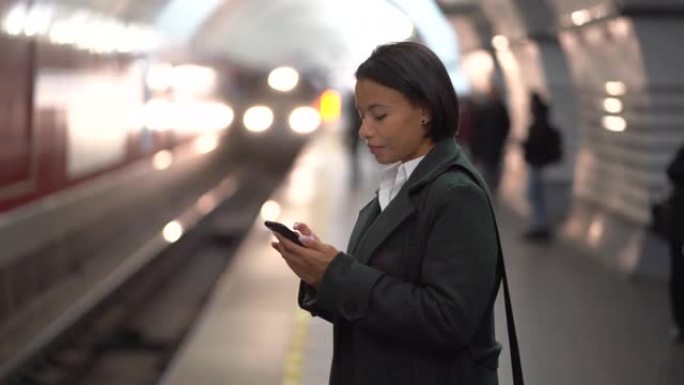 严肃的非洲女商人在等待地铁列车时在地铁站台上使用智能手机