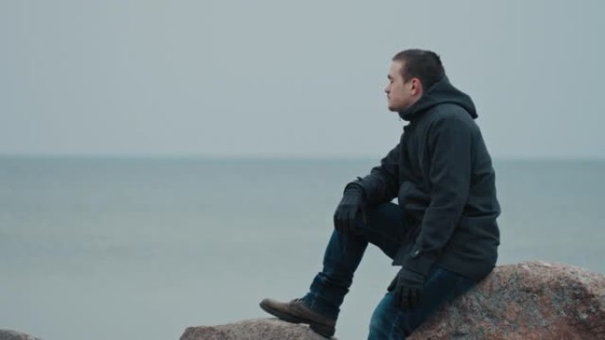 一个年轻人坐在蓝色大海的背景下的石头上，思考