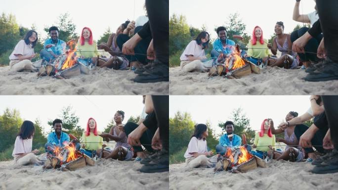 不同国籍的朋友坐在沙滩上，坐在沙滩上，喝啤酒，用火烧香肠，吃棉花糖，谈论在一起的时间，一起笑