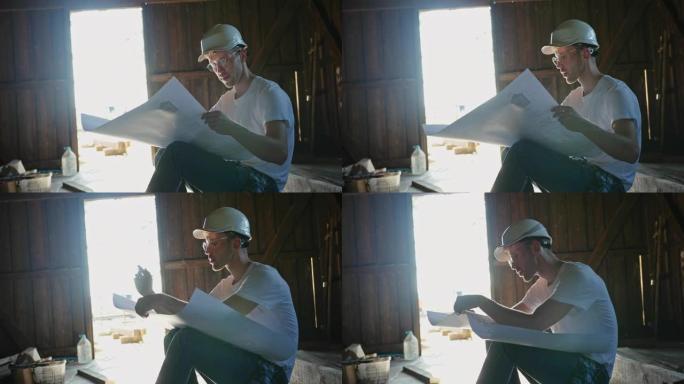 戴着安全帽的年轻男性建筑商坐在木棚里，在晴天在蓝图上做笔记
