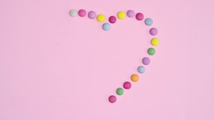 彩色彩虹糖果在明亮的粉红色背景下形成浪漫的心形。停止运动平铺概念