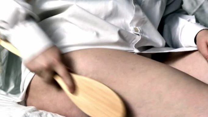 在家身体护理，脂肪治疗。一个穿着白衬衫坐在床上的女孩用按摩刷在大腿上进行自我按摩，用力摩擦问题区域