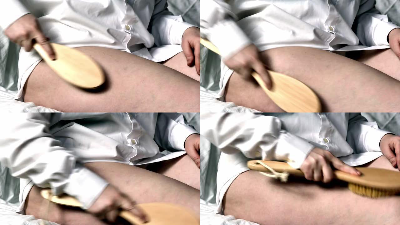 在家身体护理，脂肪治疗。一个穿着白衬衫坐在床上的女孩用按摩刷在大腿上进行自我按摩，用力摩擦问题区域