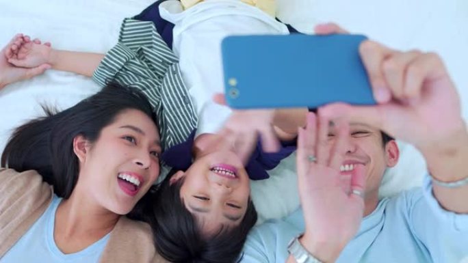 幸福的家庭时光，拿着亚洲父母的手机自拍，爸爸，妈妈和儿子一起躺在卧室的床上，开心地笑着。周末用爱养育