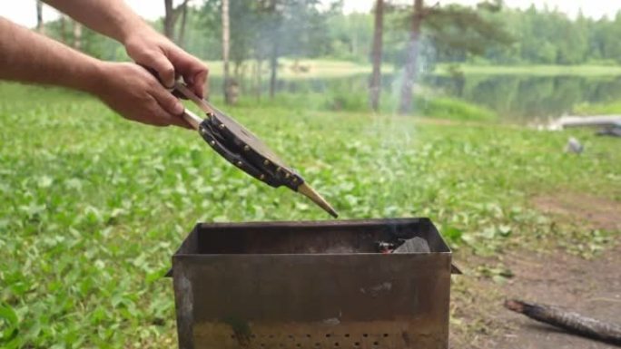 一名男子在烧烤用硬木壁炉风箱吹火。在湖边钓鱼之旅中在树林里休息