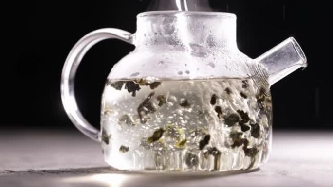 在深色背景下的玻璃透明茶壶中酿造凉茶的过程。将热水倒入带茶叶的水壶中。