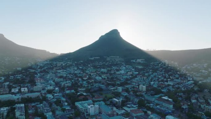 高高的尖峰在城市的住宅区投下阴影。空中上升的镜头揭示了狮子头山后面的灿烂阳光。南非开普敦