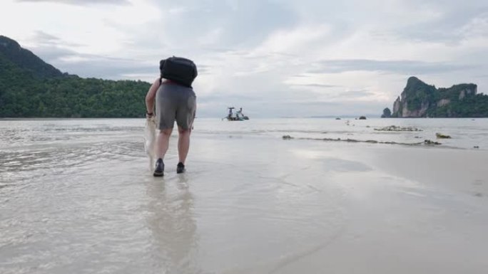 亚洲男性背包旅游志愿者手持塑料垃圾袋走进低潮海水，积极的自然环境和人，垃圾清除活动，可持续旅游