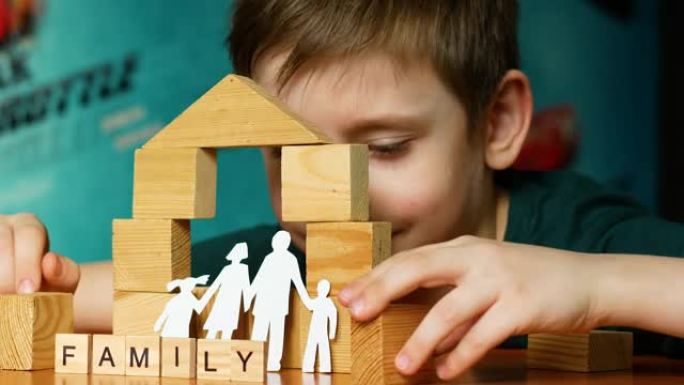 一个7岁的高加索男孩用木制立方体建造了一个玩具屋，即铭文家族，一个家庭的剪影是用纸剪掉的。孩子的梦想