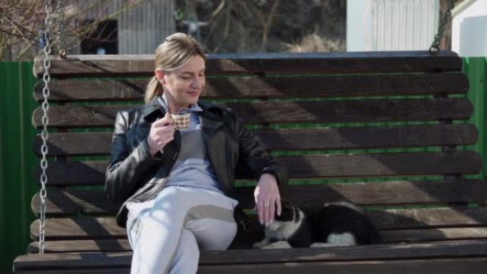 一个满意的女人坐在花园里的长椅上，轻轻地抚摸着猫。宠物护理，友谊。