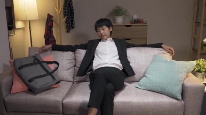 在客厅里劳累了一天的工作后，回到家中的亚洲女工在沙发坐垫和交叉腿的情况下，用乏力触摸僵硬的脖子