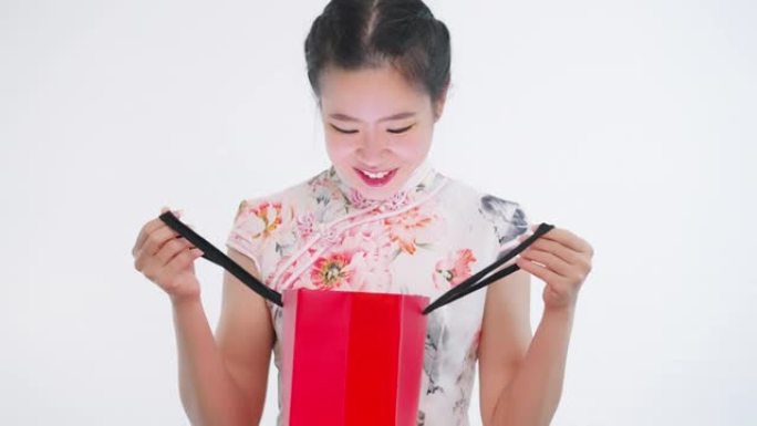 中国/亚洲妇女在春节期间打开红色购物袋迎接惊喜，白色背景，穿着旗袍白色碎花图案连衣裙