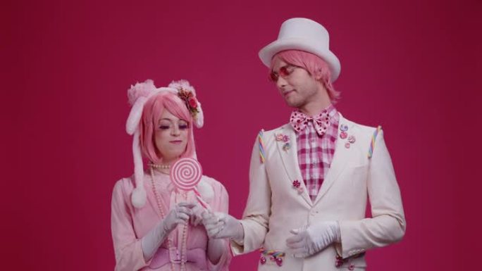几个穿着粉红色服装的彩色演员哑剧站在粉红色的工作室背景上，一个男人和一个女孩调情，给了一个棒棒糖，并