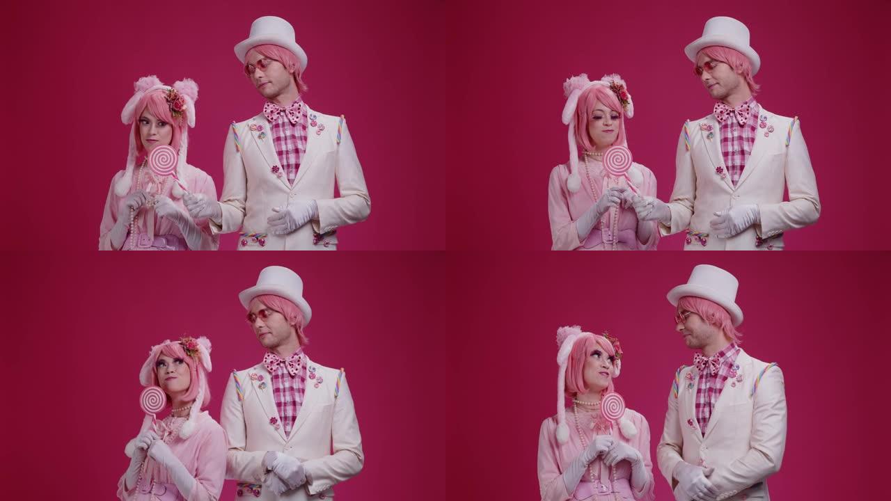 几个穿着粉红色服装的彩色演员哑剧站在粉红色的工作室背景上，一个男人和一个女孩调情，给了一个棒棒糖，并