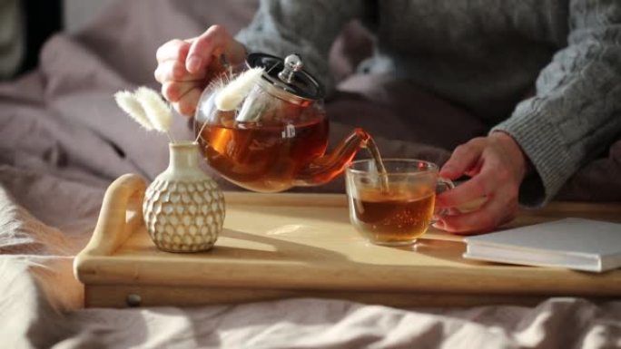 女人用玻璃茶壶在杯子里倒茶。舒适的家居hygge风格内饰。早餐时的茶道。