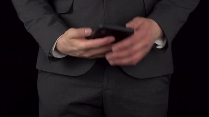 一个穿着西装的年轻商人从口袋里拿出一部电话，并在里面发短信。黑色背景上的一个男人的腰部特写。