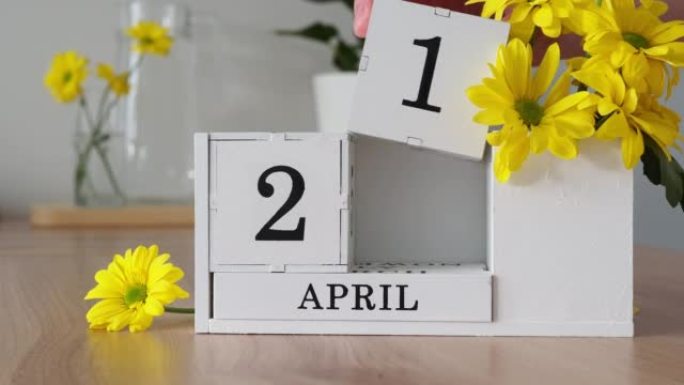 春季月份4月21日。女人的手翻过一个立方历法。黄色花朵旁边的桌子上的白色万年历。在一个月内更改日期。