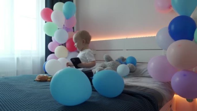 可爱的小孩在卧室的床上玩智能手机，上面装饰着五颜六色的气球，1岁的男婴在家庆祝生日。