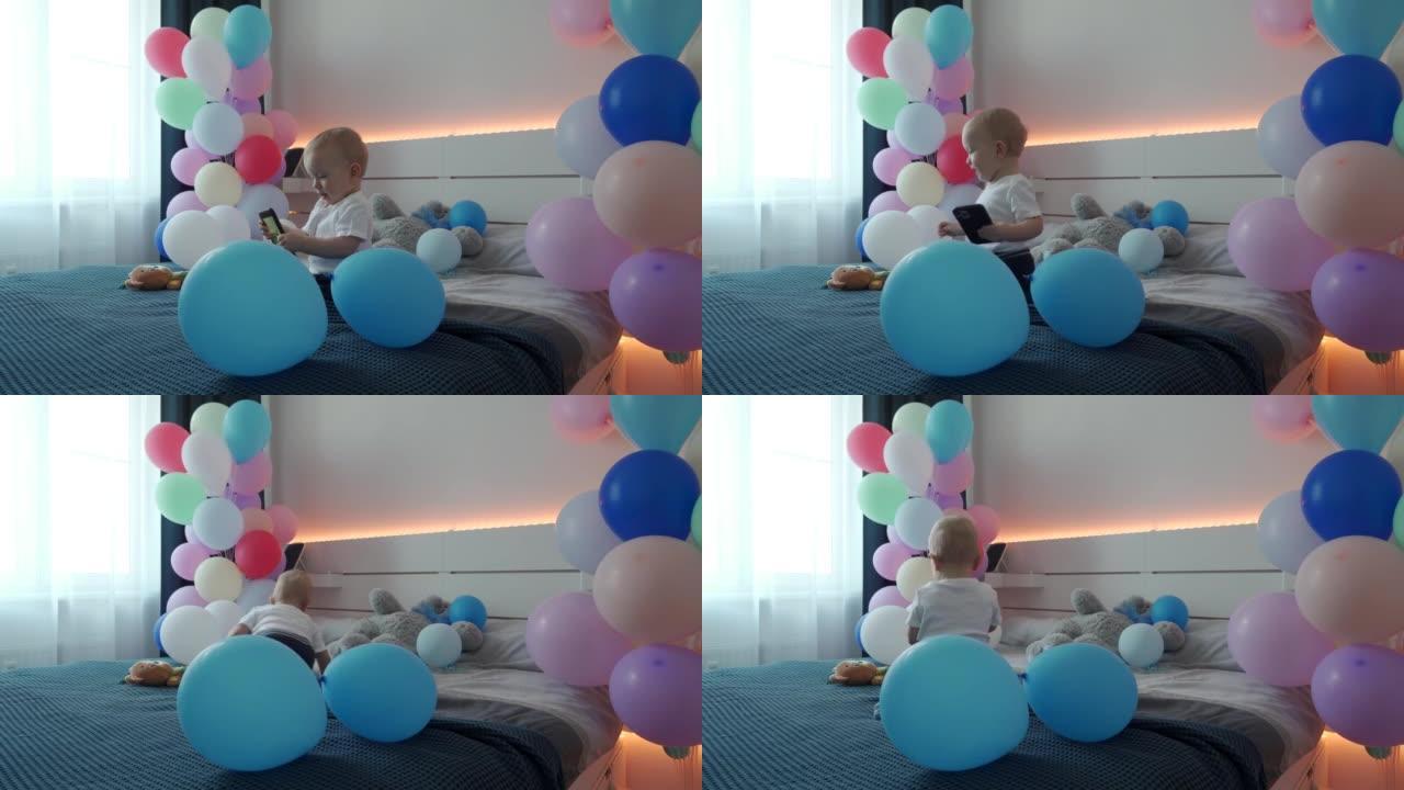 可爱的小孩在卧室的床上玩智能手机，上面装饰着五颜六色的气球，1岁的男婴在家庆祝生日。