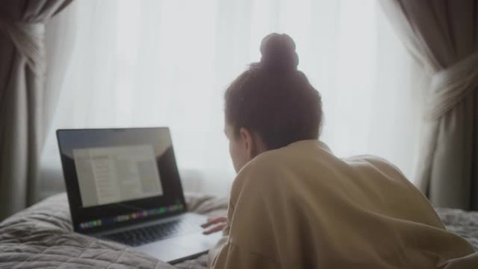 集中的女人在笔记本电脑上工作。女性自由职业者使用现代PC。女士拿着笔记本躺在床上。女性博客作者家庭工