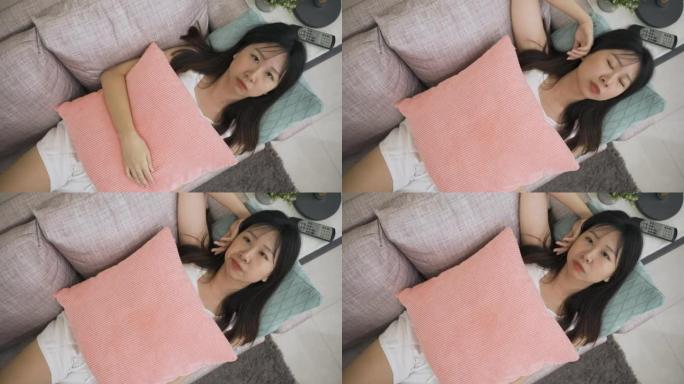 高角度拍摄懒惰的亚洲女人凝视着天花板，躺在家里的沙发上时，她的手叹息着脸，感觉无所事事。