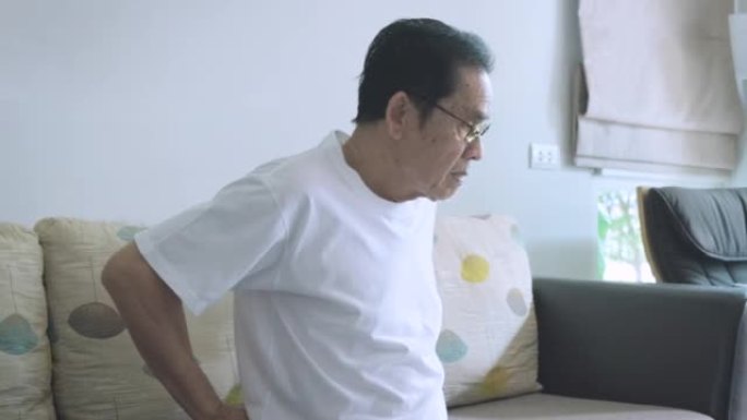亚洲老人带一只手到腰上缓解背部疼痛，老人背痛在客厅家沙发上靠窗