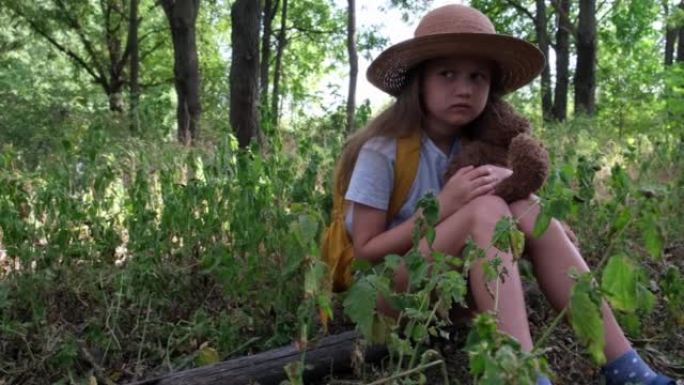 带着泰迪熊和黄色背包的小女孩独自坐在森林里。儿童游客迷失在灌木丛中。心烦意乱的孩子拥抱一个玩具，等待