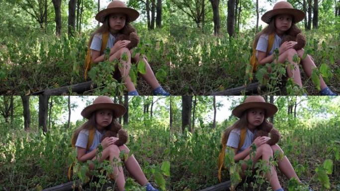 带着泰迪熊和黄色背包的小女孩独自坐在森林里。儿童游客迷失在灌木丛中。心烦意乱的孩子拥抱一个玩具，等待