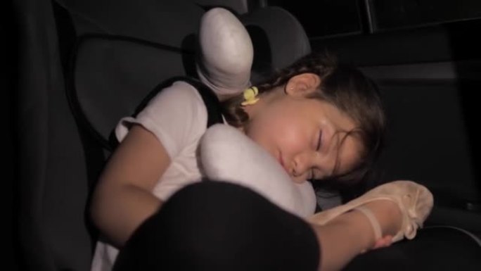 一个小女孩晚上睡在汽车座椅的后座上。汽车在路上。开车长途旅行后，孩子累了。不舒服的睡姿。自动睡觉枕头