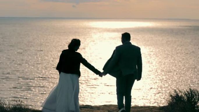 穿着便服的新婚夫妇在海边走向夕阳。新娘和新郎散步，手牵手，拥抱日落时的海洋全景。婚礼的概念