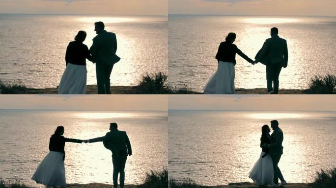 穿着便服的新婚夫妇在海边走向夕阳。新娘和新郎散步，手牵手，拥抱日落时的海洋全景。婚礼的概念