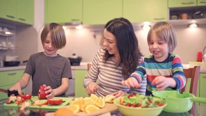 幸福的年轻家庭，美丽的母亲和两个孩子，可爱的学龄前男孩和在校男孩在阳光明媚的厨房里一起做饭。