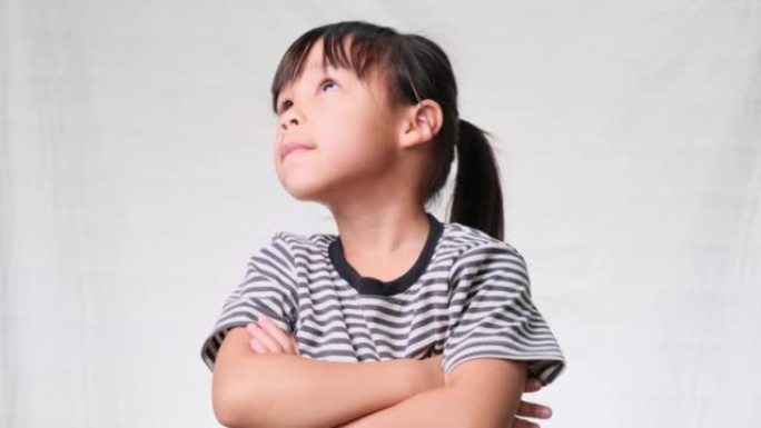 可爱的小女孩双臂交叉抬头，在白色工作室背景下思考一些事情。想象的概念