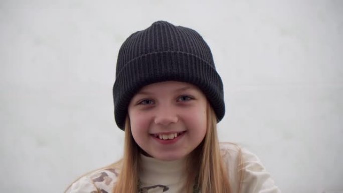 开朗的积极向上的学龄女孩，戴着白色背景上的温暖帽子。她看着镜头时微笑着。