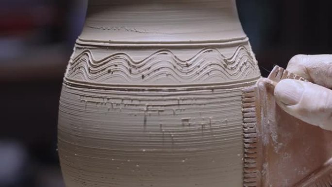 陶工的男性手使用陶器工具在柔软的白色陶罐上雕刻图案。陶瓷器皿在创意车间的陶工轮上旋转。陶器在柔和的光