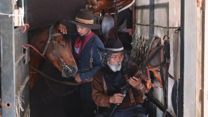 两个穿着牛仔服装的男人呆在马stable里，一个检查长枪的子弹，另一个在后面照顾马
