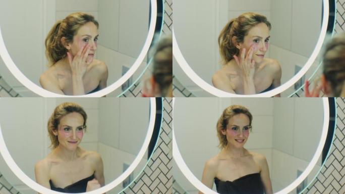 美丽的女人在浴室镜子附近涂眼罩。年轻迷人的女人用手指敲击眼睛。